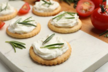 Krem peynirli krakerler, biberiye ve domatesler beyaz masada, yakın plan.