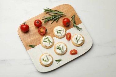 Krem peynirli krakerler, biberiye ve domatesler beyaz masada.