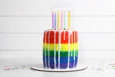 Lezzetli gökkuşağı pastası yanan mumlar ve beyaz ahşap zemin üzerine serpiştirilmiş şekerlemeler.