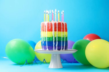 Nefis gökkuşağı pastası yanan mumlar ve açık mavi arka planda balonlar