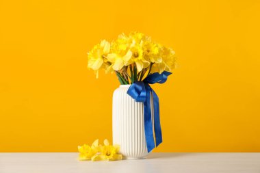 Beyaz masada sarı arka planda vazoda kurdeleli güzel nergisler.