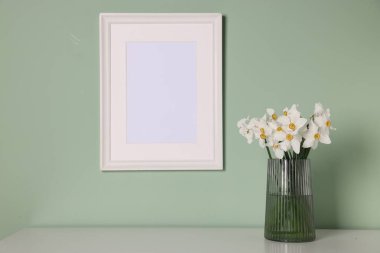 Vazodaki güzel nergisler ve resim çerçevesi açık yeşil duvarın yanındaki beyaz masada.