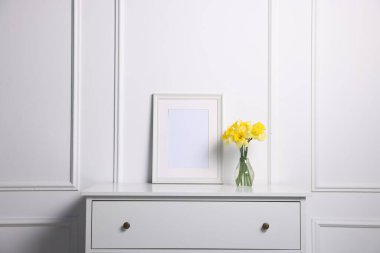 Evdeki beyaz duvarın yanındaki dolapta vazoda ve resim çerçevesinde güzel nergisler var.