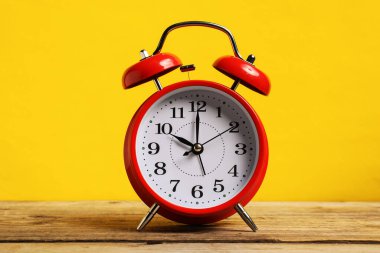 Sarı arka planda, ahşap masada kırmızı bir çalar saat.