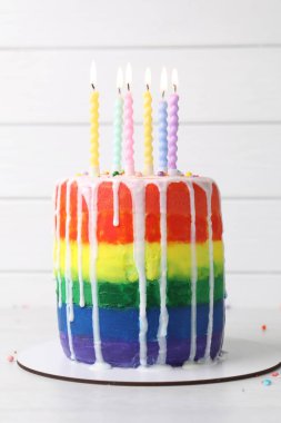 Lezzetli gökkuşağı pastası, yanan mumlar ve beyaz masanın üzerine serpiştirilmiş şekerler.