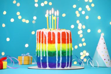 Enfes gökkuşağı doğum günü pastası, yanan mumlar, flamalar, hediye kutuları ve açık mavi masa üzerindeki parti şapkası bulanık ışıklara karşı, yakın çekim