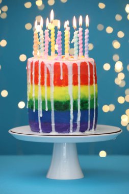 Bulanık ışıklara karşı açık mavi masada yanan mumlarla nefis gökkuşağı pastası, yakın plan.