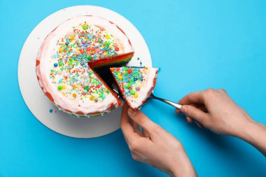 Bir kadın açık mavi masada spatulayla lezzetli bir gökkuşağı pastası yiyor.