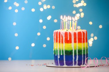 Enfes gökkuşağı doğum günü pastası, yanan mumlar, serpiştirmeler ve mavi arka plandaki ışık paneli, bulanık ışıklar, yakın plan. Metin için boşluk
