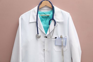 Doktor ceketi, steteskop ve rozeti bej arka planda asılı.