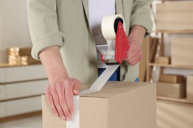 Karton kutuyu kapalı bir yere kapatmak için yapıştırıcılı bant kullanan bir adam.