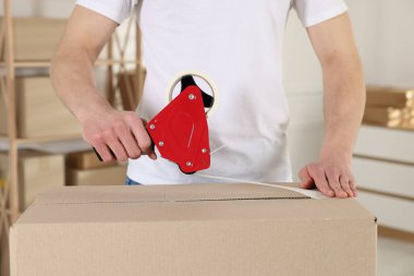 Karton kutuyu kapalı bir yere kapatmak için yapıştırıcılı bant kullanan bir adam.