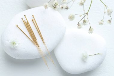 Akupunktur iğneleri, çakıl taşları ve çiçekler beyaz masada, düz yerde.