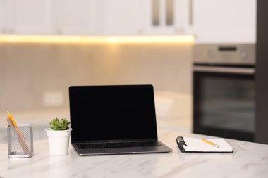 Boş ekranlı dizüstü bilgisayar, mutfaktaki beyaz mermer masada ev bitkisi ve kırtasiye malzemesi. Tasarım için model