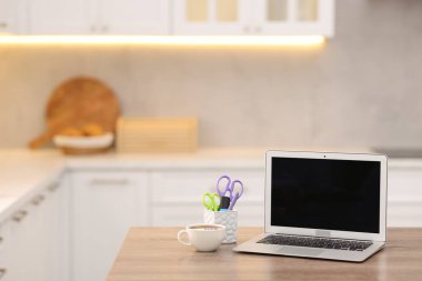 Boş ekranlı dizüstü bilgisayar, mutfaktaki ahşap masada bir fincan sıcak içecek ve kırtasiye malzemesi. Tasarım için model