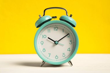 Sarı arka planda beyaz ahşap masada bir turkuaz alarm saati.