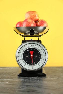 Mekanik mutfak terazisi, sarı arka planda koyu dokuma masa üzerinde elma kasesi.