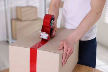 Karton kutuyu kapalı bir yere kapatmak için yapıştırıcılı bant kullanan bir adam.