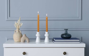 Yakıcı mumları olan şık şamdanlar vazoda kuru bitkiler ve kapalı alandaki gri duvarın yanındaki beyaz çekmecelerin üzerindeki diğer dekorlar.