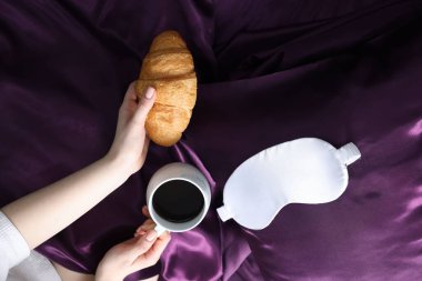 Kahve, kruvasan ve uyku maskesiyle mor ipek çarşaflı bir kadın.