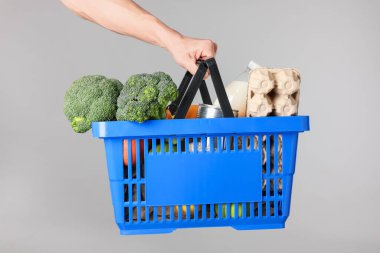 Elinde gri arka planda farklı ürünler olan alışveriş sepeti tutan adam, yakın plan.