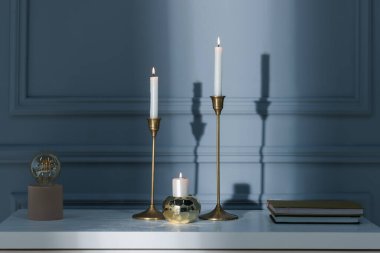 İçinde yanan mumlar, kitaplar ve lambalar olan şık şamdanlar kapalı alandaki gri duvarın yanındaki beyaz masada.