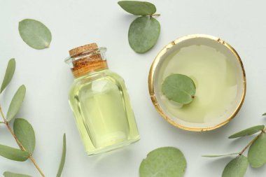 Beyaz arka planda bir şişe temel yağ, kase ve okaliptüs yaprakları, düz uzanır.