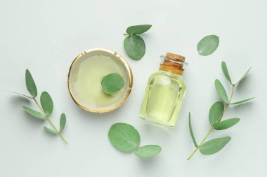 Beyaz arka planda bir şişe temel yağ, kase ve okaliptüs yaprakları, düz uzanır.