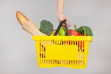 Elinde gri arka planda farklı ürünler olan alışveriş sepeti tutan adam, yakın plan.