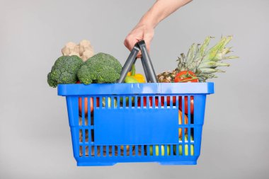 Elinde gri arka planda farklı ürünler olan alışveriş sepeti tutan adam, yakın plan.