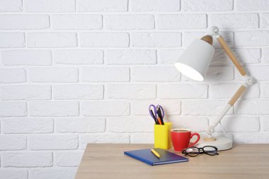 Farklı kırtasiye malzemeleri, kahve, lamba ve bardaklar beyaz tuğla duvarın yanındaki ahşap ofis masasında.