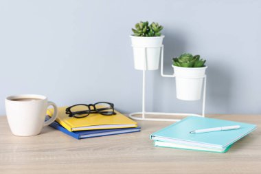 Farklı kırtasiye malzemeleri, bardaklar, kahve ve çiçeklerle aydınlık duvarın yanındaki ahşap ofis masasında.