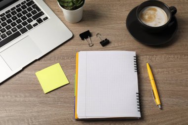 Not defteri, kalem, yapışkan not, cilt klipleri, kahve, çiçek dekoru ve dizüstü bilgisayar ahşap masanın üstünde.