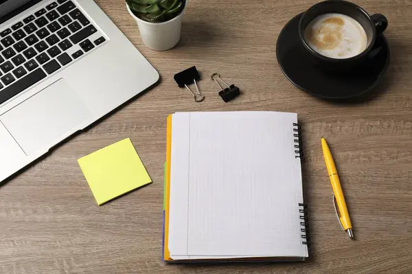Not defteri, kalem, yapışkan not, cilt klipleri, kahve, çiçek dekoru ve dizüstü bilgisayar ahşap masanın üstünde.