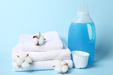 Bir şişe yumuşatıcı, pamuk çiçekleri ve açık mavi arka planda havlular.