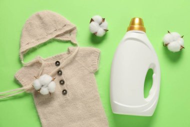 Bir şişe yumuşatıcı, pamuk çiçekleri ve yeşil arka planda bebek kıyafetleri, düz uzanmış.