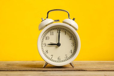 Ahşap masada sarı arka planda beyaz bir çalar saat.