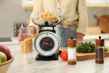 Kadın, mekanik mutfak terazisinin yanında hamur makarna kabı tutuyor. Beyaz mermer tezgahta, yakın plan.