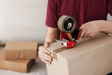 Karton kutuyu kapalı bir yere kapatmak için yapıştırıcılı bant kullanan bir adam.