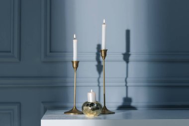Gri duvarın yanındaki beyaz masada yanan mumlarla süslü şamdanlar.