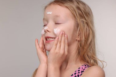 Gülümseyen küçük kız, gri arka planda yüzüne güneş kremi sürüyor.