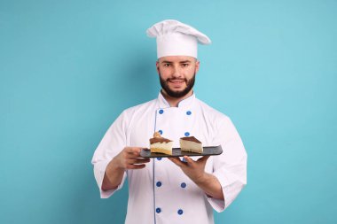 Üniformalı mutlu pastacı elinde açık mavi arka planda lezzetli peynirli kek parçaları tutuyor.
