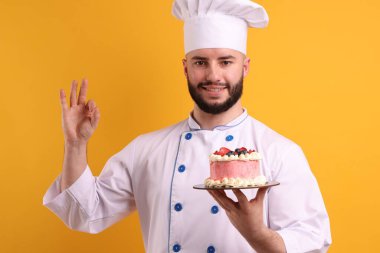 Üniformalı mutlu pastacı elinde böğürtlenlerle turuncu arka planda iyi niyet gösterisi yapıyor.