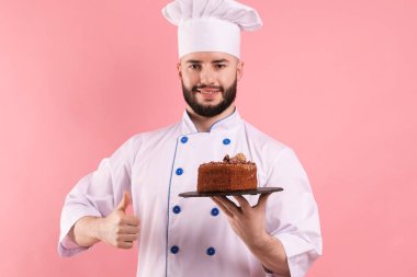 Üniformalı mutlu pastacı elinde çikolatalı pasta ve pembe arka planda baş parmak gösteriyor.