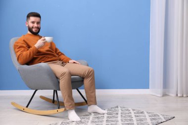 Elinde bir bardak sıcak içecekle gülümseyen adam evdeki sallanan koltukta dinleniyor. Metin için boşluk