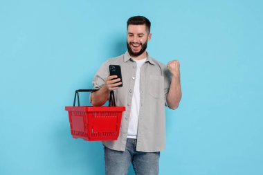 Boş alışveriş sepeti ve açık mavi arka planda akıllı telefonu olan bir adam.