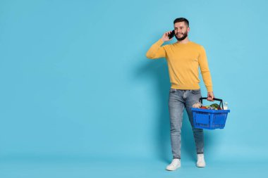 Alış veriş sepeti dolusu ürünü olan bir adam akıllı telefondan açık mavi arka plana karşı konuşuyor. Mesaj için yer var.