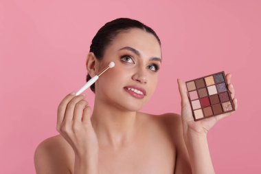 Göz farı paleti ve fırçası olan pembe arka planda makyaj yapan kadın.