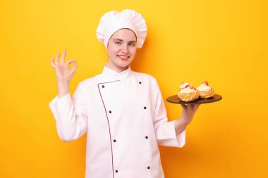 Üniformalı bir şekerci lezzetli profiteroller tutuyor ve turuncu arka planda iyi bir jest yapıyor.