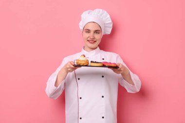 Üniformalı şekerci pembe arka planda farklı lezzetli tatlılar tutuyor.